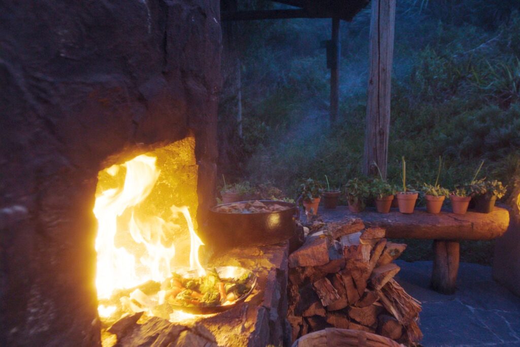 Stone outdoor hearth with a cast-iron pot and pan over a wood fire at dusk on a New Zealand property